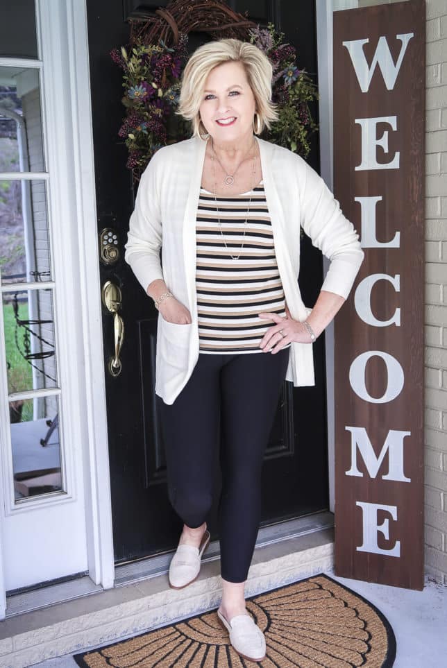 Fashion Blogger 50 Is Not Old wearing a striped tank top, black leggings, a white long cardigan, and linen slides