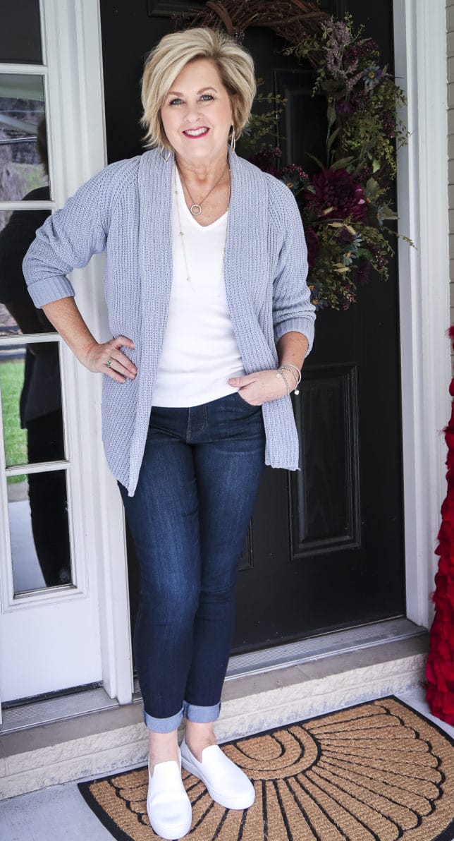 Fashion Blogger 50 Is Not Old is wearing a white v-neck t-shirt, a blue chunky knit cardigan, dark wash jeggings, and white slip-on slippers