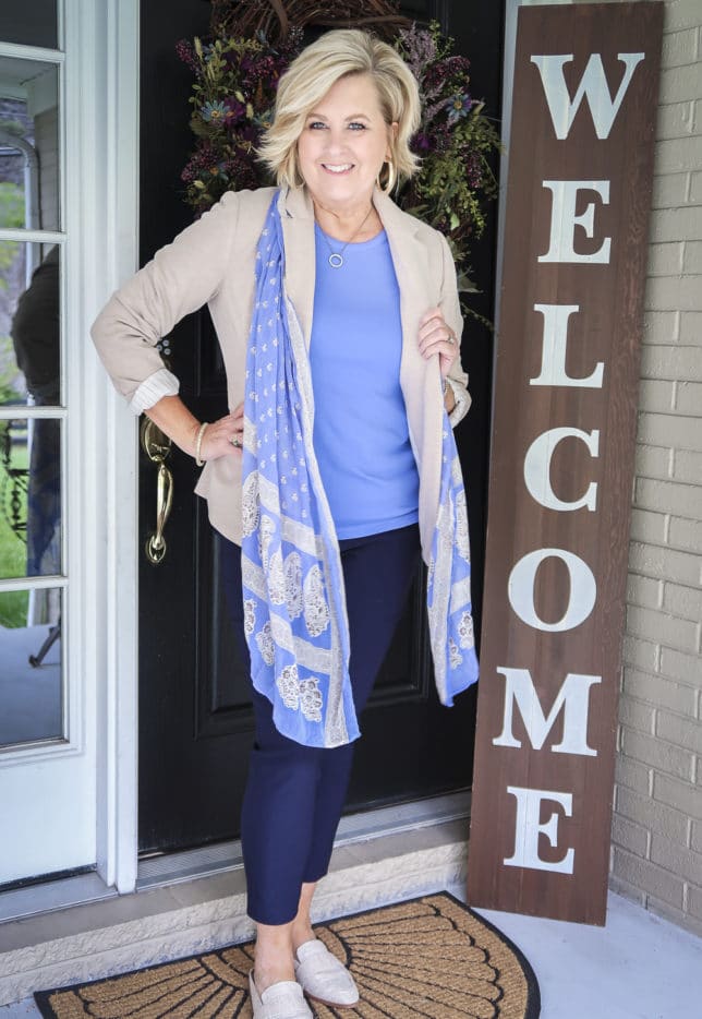 Fashion Blogger 50 Is Not Old is wearing a periwinkle blue t-shirt with navy ankle pants, a beige blazer, a blue scarf, and linen looking mules