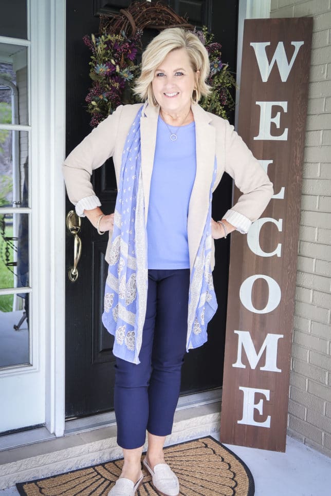 Fashion Blogger 50 Is Not Old is wearing a periwinkle blue t-shirt with navy ankle pants, a beige blazer, a long blue scarf, and linen looking mules