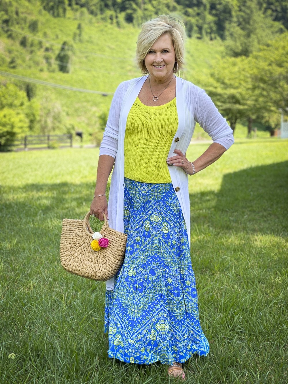 Fashion Blogger 50 Is Not Old is wearing styles from Walmart. A long white cardigan, a lemon yellow sweater tank top, a blue tiered skirt, embellished sandals, and a straw handbag