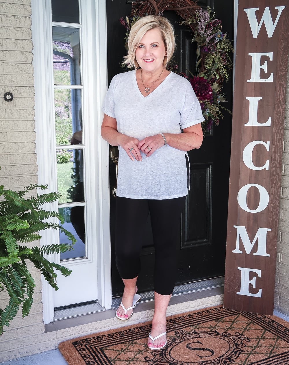 FASHION BLOGGER 50 IS NOT OLD IS WEARING A LIGHT GRAY TUNIC TOP AND BLACK CAPRI LEGGINGS WITH A PAIR OF WHITE FLIP FLOPS