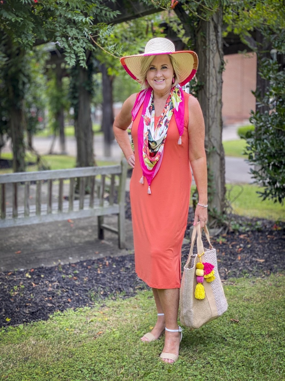 Fashion Blogger 50 Is Not Old is wearing an orange midi dress with a tropical print scarf in pink, a yarn trimmed straw hat, and carrying a jute tote with colorful pompoms all from Chico's