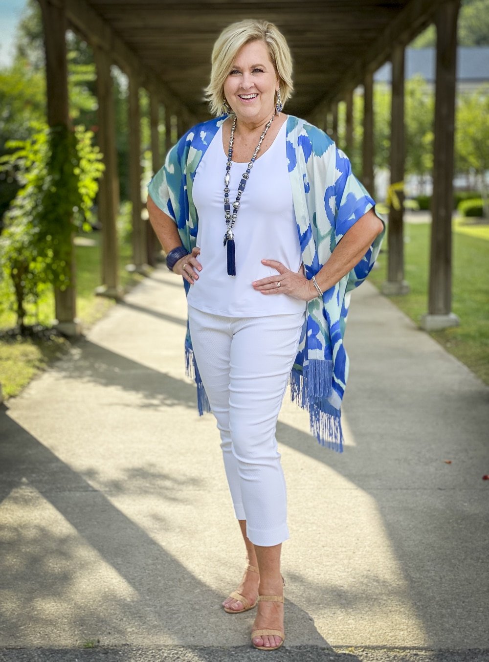 Fashion Blogger 50 Is Not Old is wearing a white tank top, white textured crop pants, and a colorful silk ruana from Chicos with a long navy necklace and bamboo heels