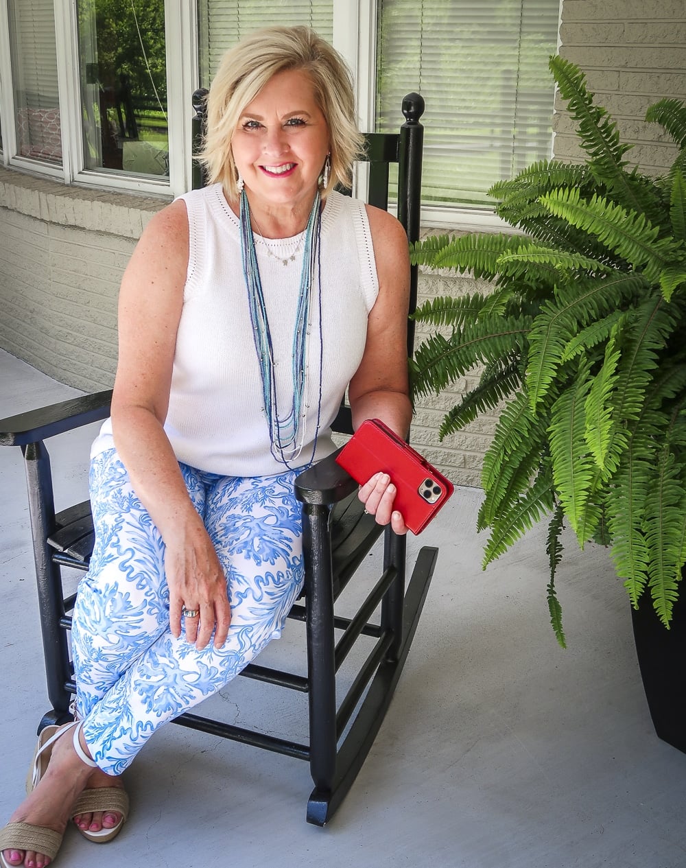 Fashion Blogger 50 Is Not Old is wearing a classic blue and white outfit sitting in a rocking chair. A white sweater, blue and white printed ankle pants from Lilly Pulitzer and espadrille wedges