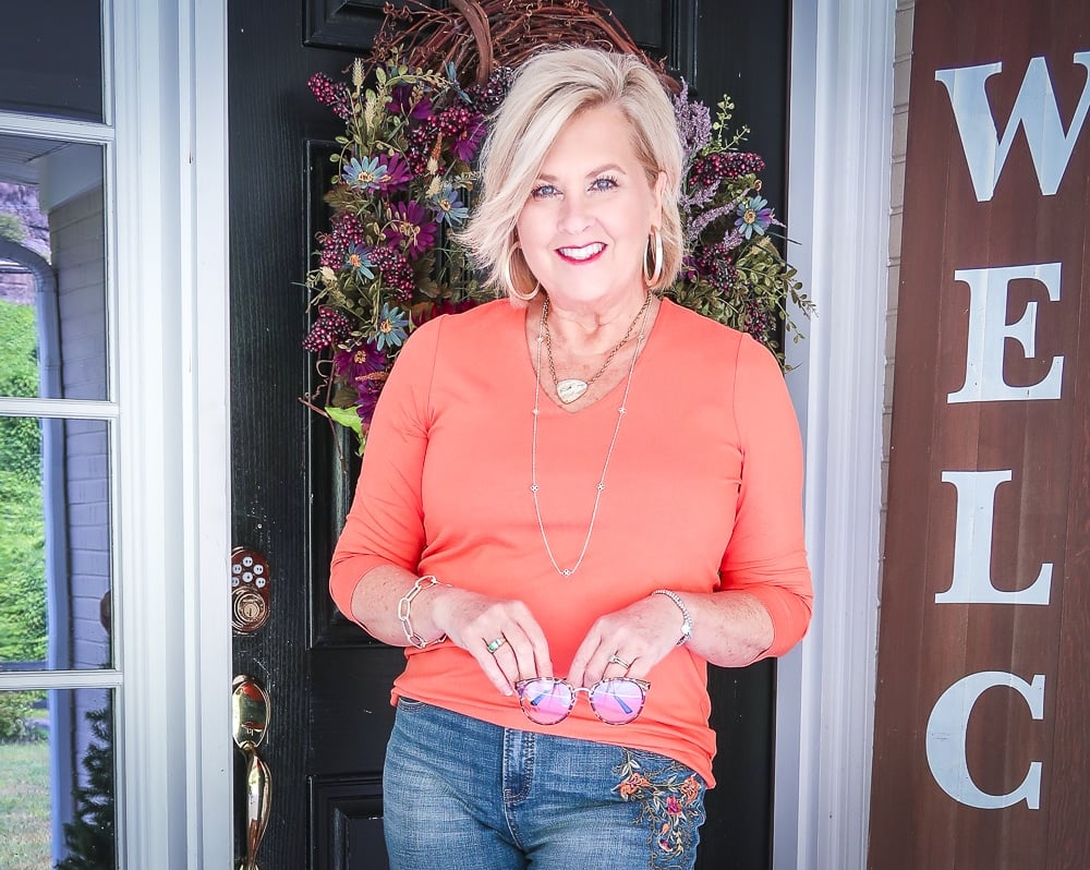 Fashion Blogger 50 Is Not Old is holding a pair of blue light blocking glasses and wearing a rust orange v-neck top from Chicos