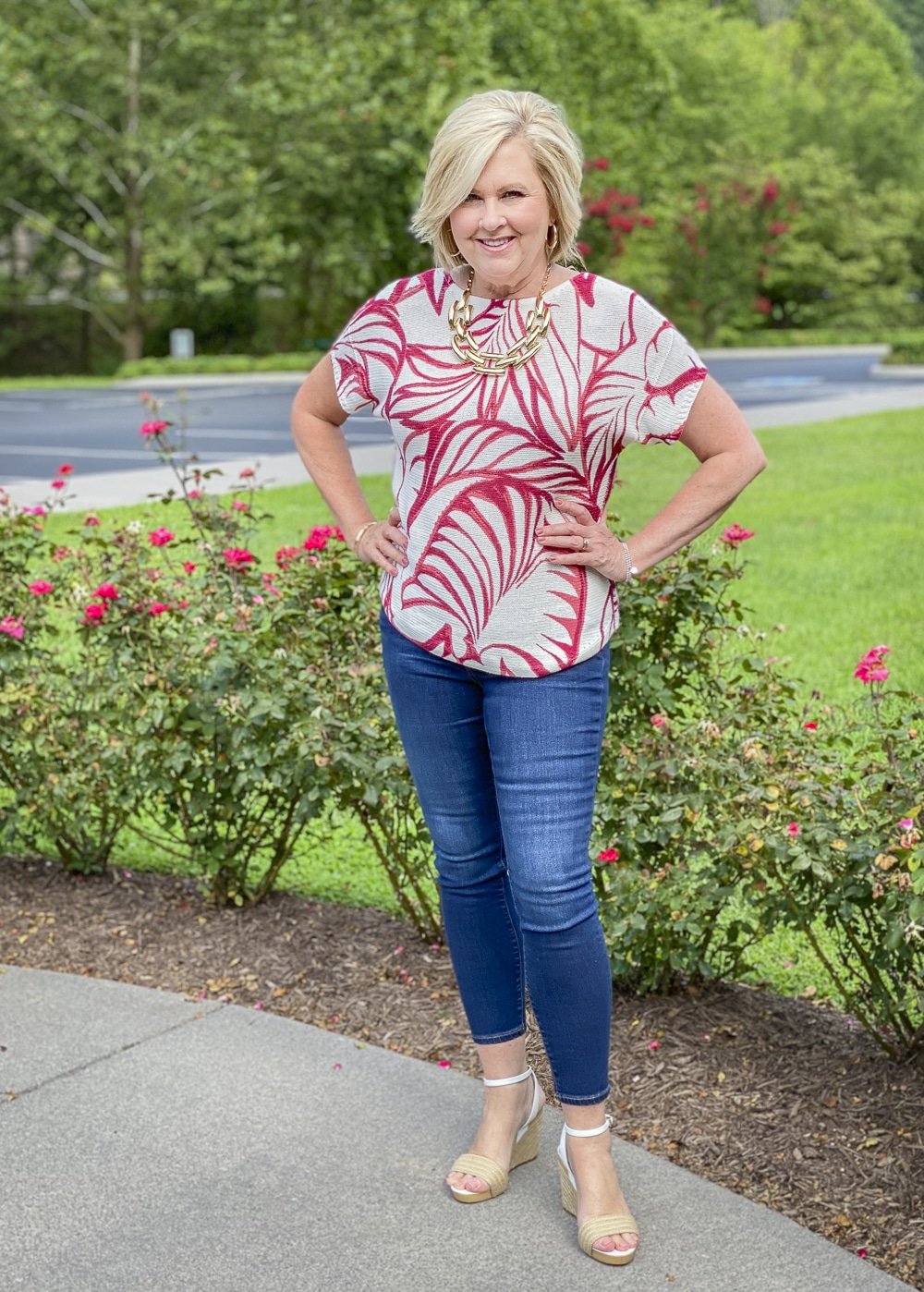 Fashion Blogger 50 Is Not Old is surrounded by red roses in her red and white tropical leaf print sweater and skinny ankle jeans from Chico's