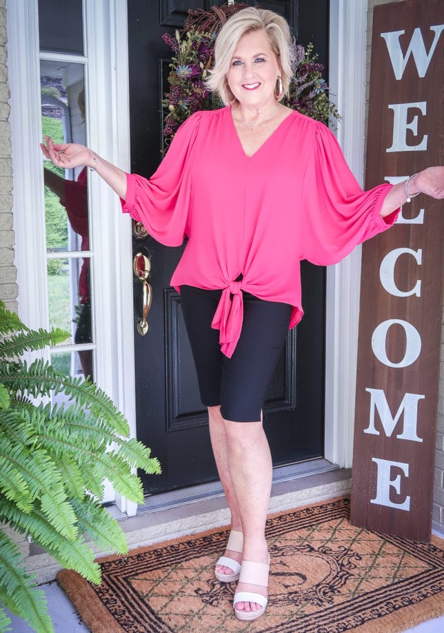 Fashion Blogger 50 Is Not Old is wearing a pink front-tie blouse and black Bermuda shorts