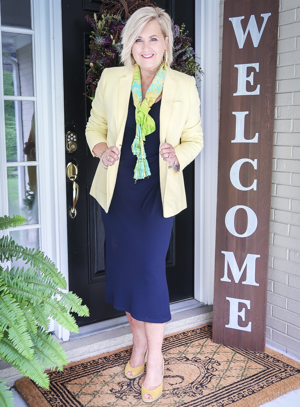 Fashion Blogger 50 Is Not Old is wearing a navy midi dress with a pale lemon yellow blazer, a bright scarf, and a pair of yellow espadrille wedge shoes