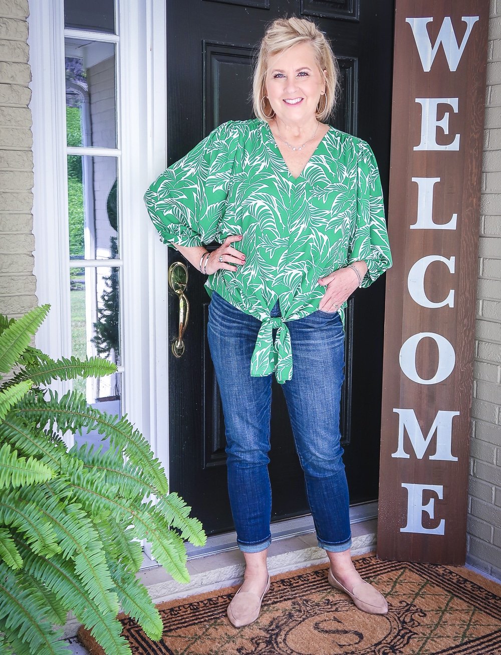 Fashion Blogger 50 Is Not Old has her hand in her pocket and is wearing a green palm print blouse and dark indigo girlfriend jeans with camel loafers