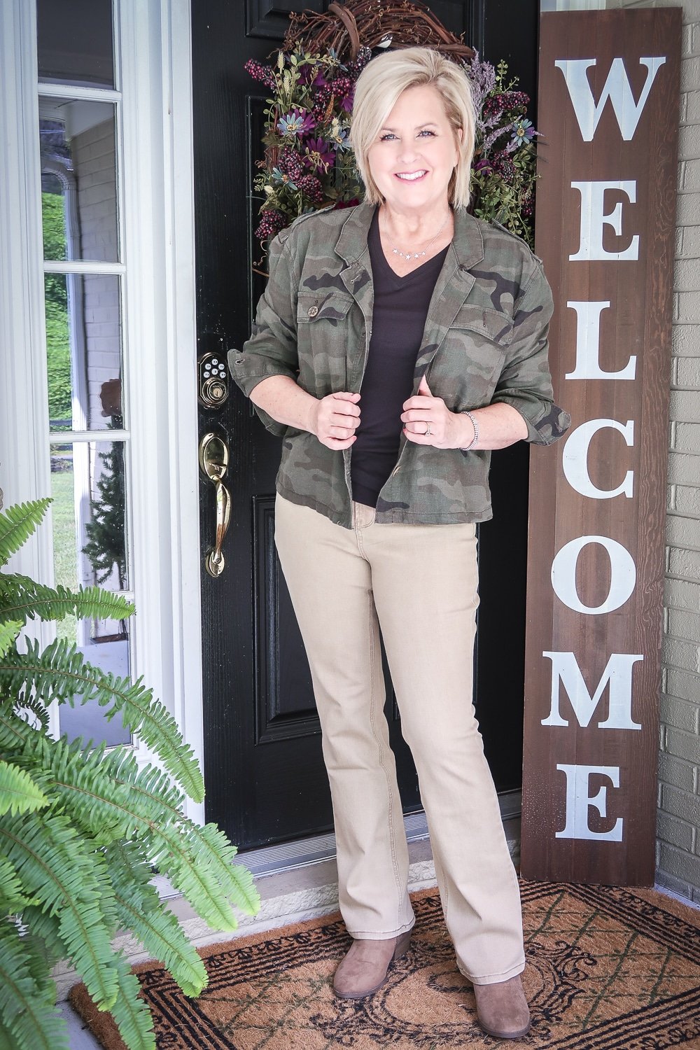 Fashion Blogger 50 Is Not Old is transitioning to fall in a black v-neck tank top, a olive green camo jacket, beige bootcut jeans, and taupe ankle booties all from Walmart