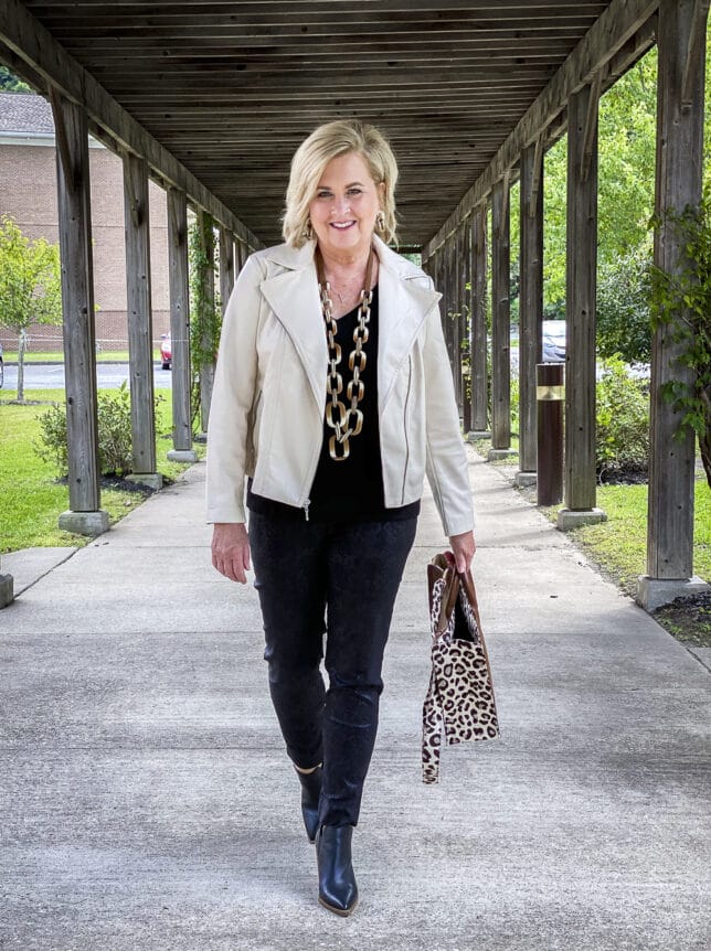 Fashion Blogger 50 Is Not Old is wearing a black top with a pair of black jeggings, a cream moto jacket, and carrying a leopard print tote 