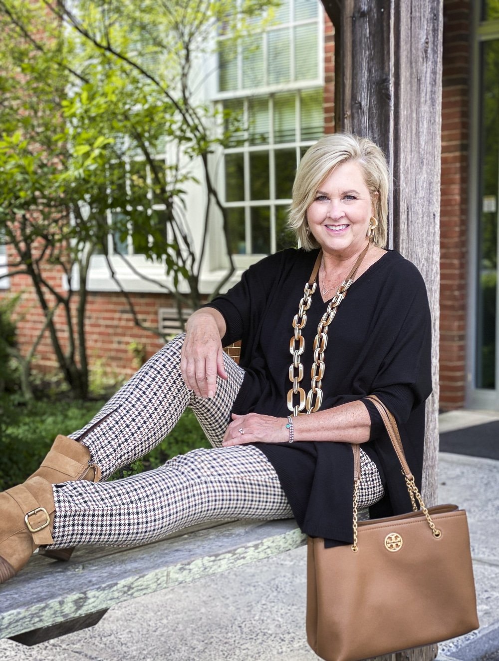 Fashion Blogger 50 Is Not Old is sitting on a bench and wearing plaid pants with a black poncho and taupe ankle boots