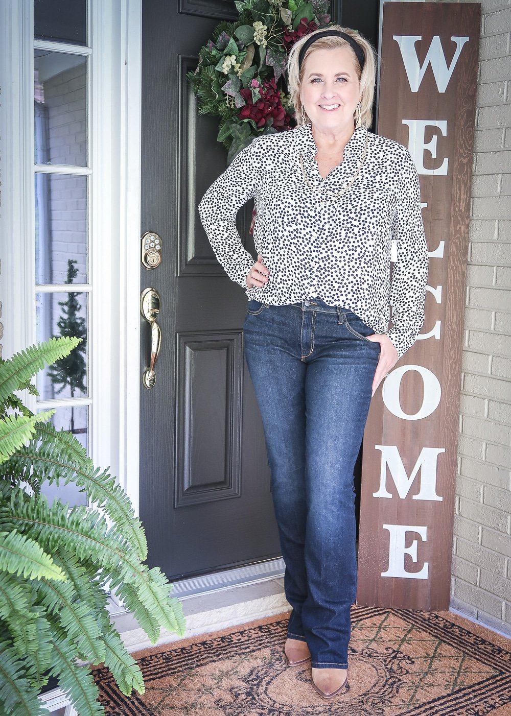 Fashion Blogger 50 Is Not Old has her hair pulled back in a headband, and wearing a dotted blouse, bootcut jeans, and ankle boots for fall