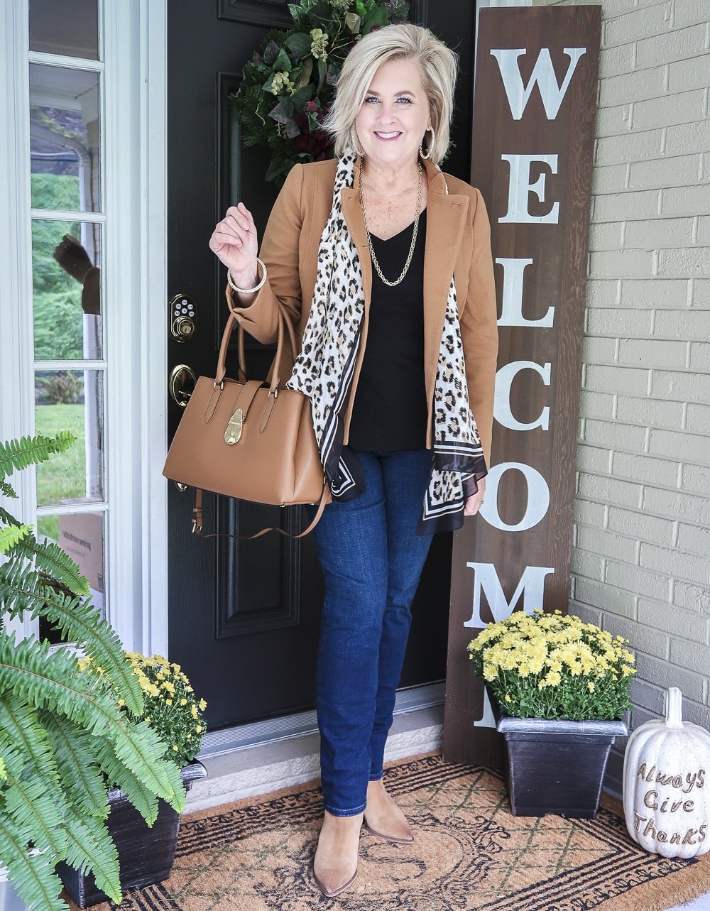 Fashion Blogger 50 Is Not Old is wearing a Camel jacket, a leopard print scarf, and black v-neck top, straight leg jeans, ankle boots, and carrying a Calvin Klein satchel tote