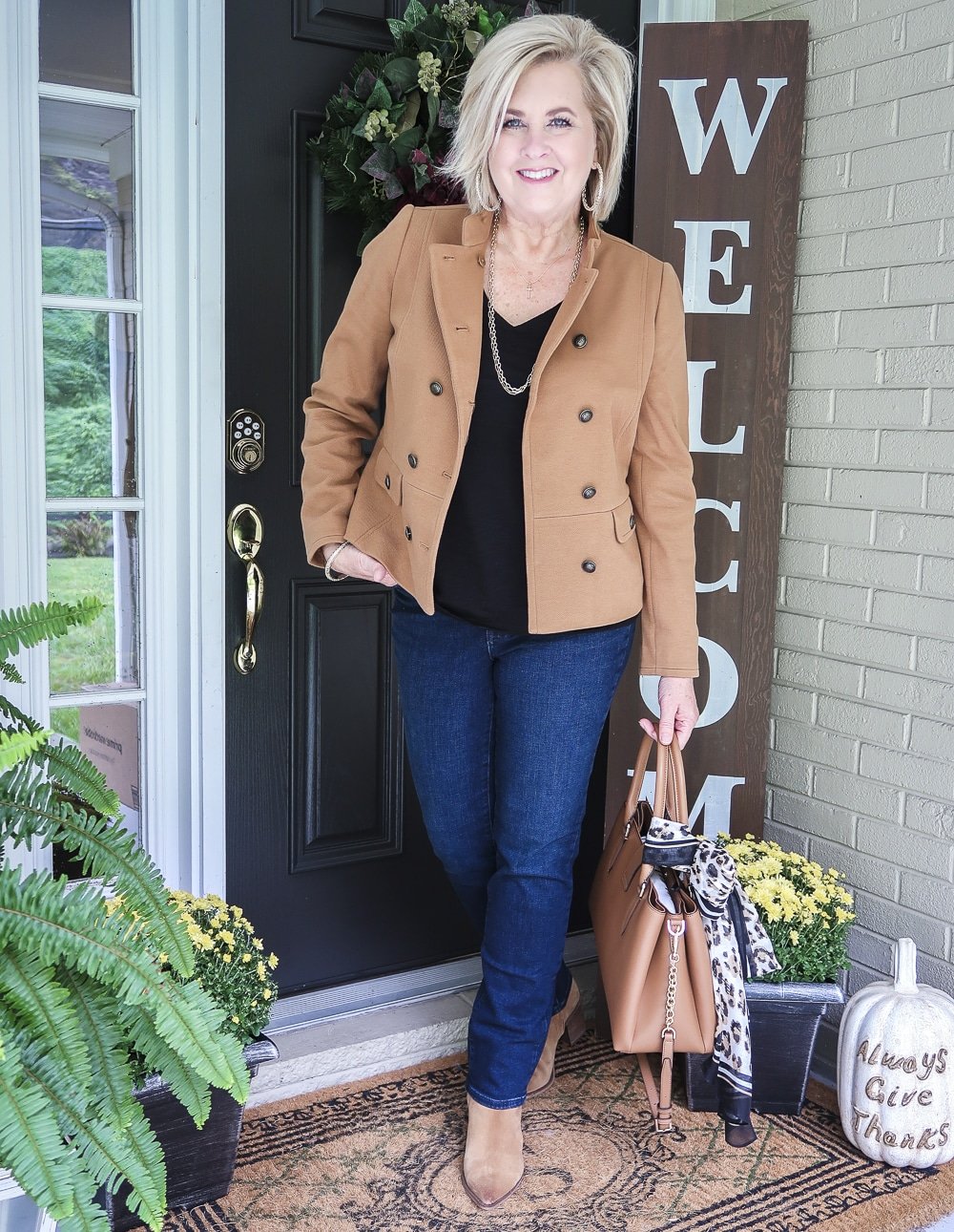 Fashion Blogger 50 Is Not Old is wearing a Camel jacket, a black v-neck top, straight leg jeans, ankle boots, and carrying a Calvin Klein satchel tote