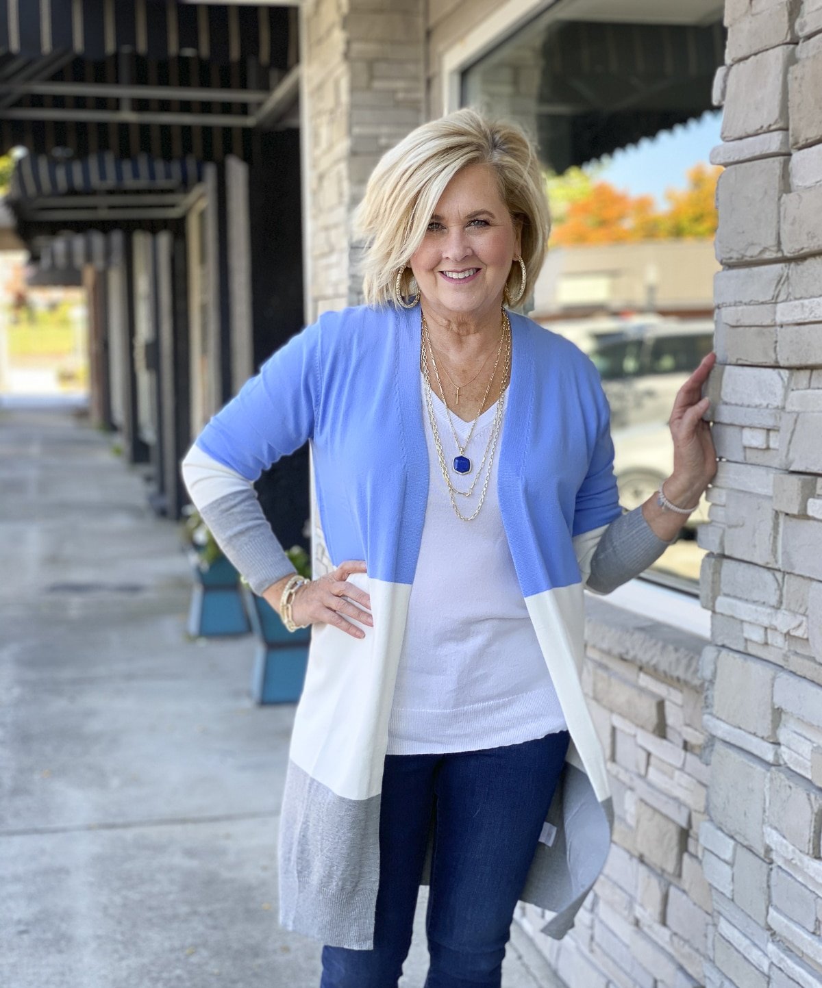 Fashion Blogger 50 Is Not Old is standing on a sidewalk in a color block sweater cardigan and leaning on a stone wall