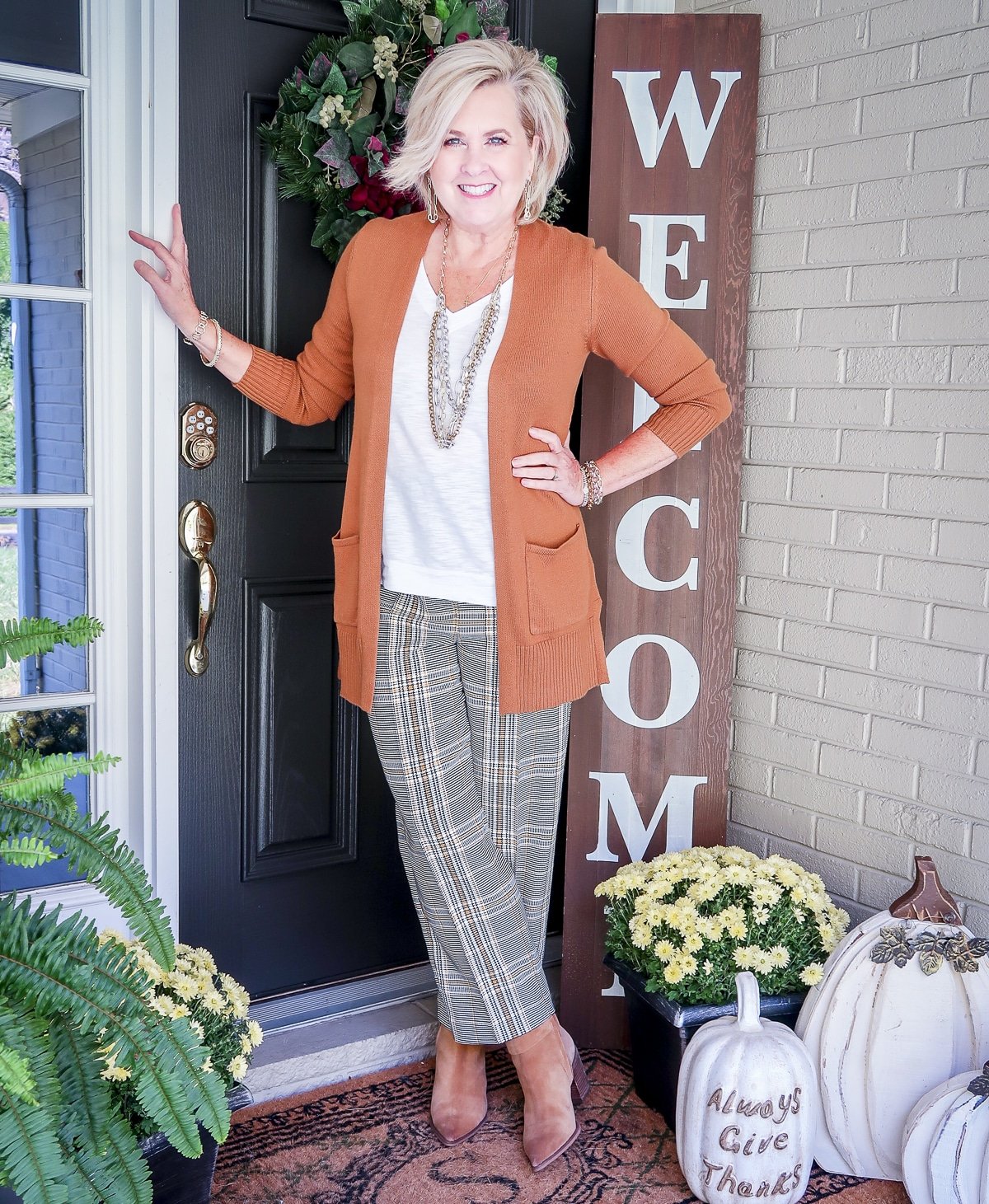 Fashion Blogger 50 Is Not Old is standing with her legs crossed in front of a black door wearing a white tank with a caramel cardigan, plaid ankle pants, and Sam Edelman ankle boots