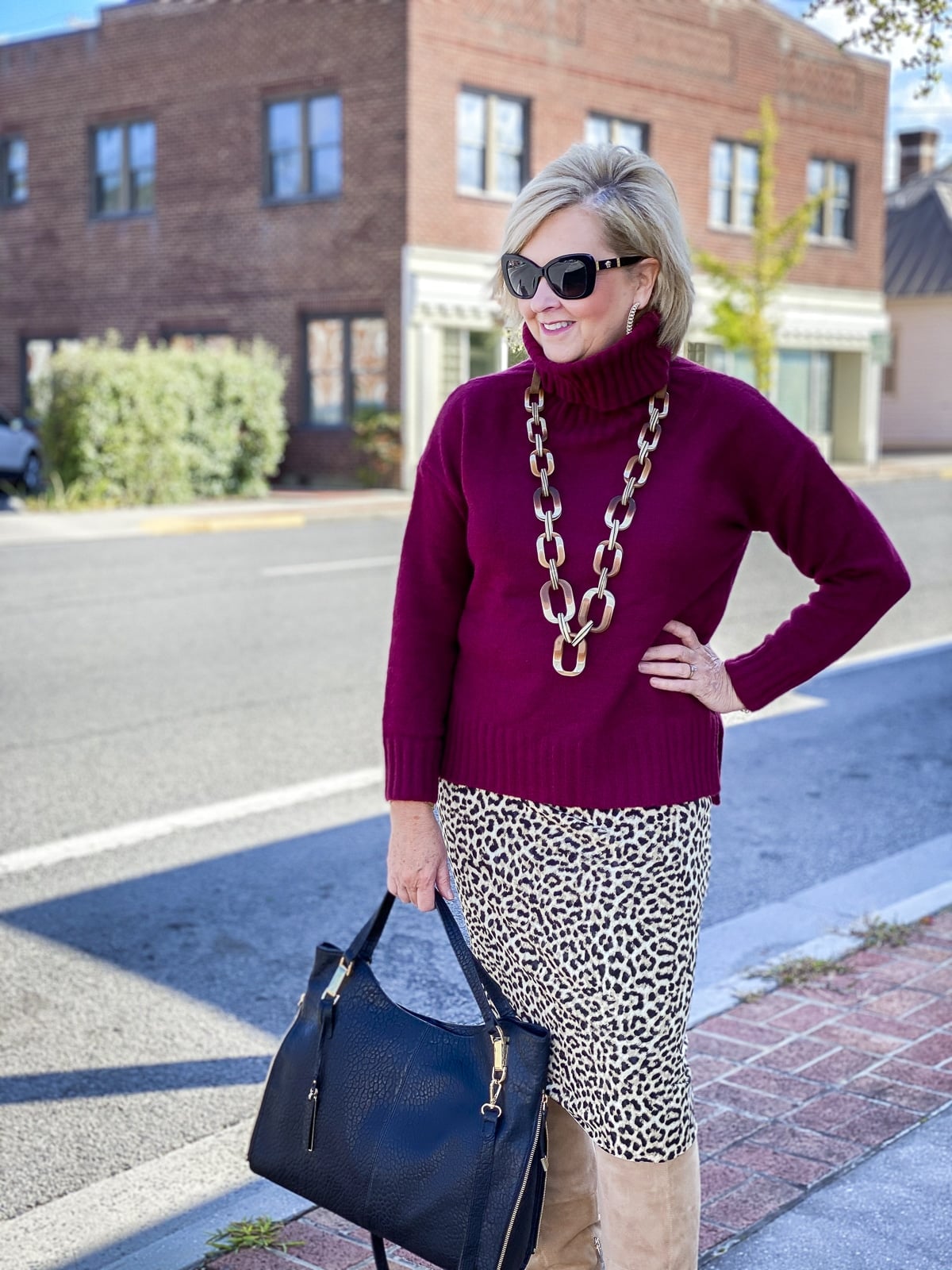 Fashion Blogger 50 Is Not Old is wearing a maroon turtleneck sweater, a pair of Versace sunglasses, a leopard tube skirt, and suede knee boots
