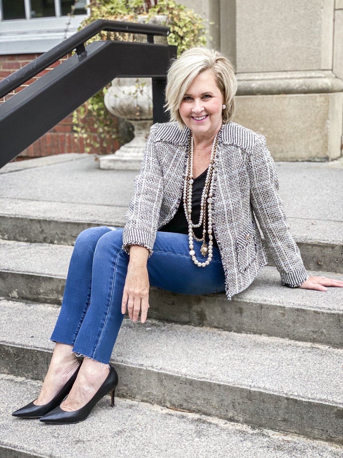 Fashion Blogger 50 Is Not Old is sitting on steps in a classic tweed blazer, distressed jeans, heels, and pearls