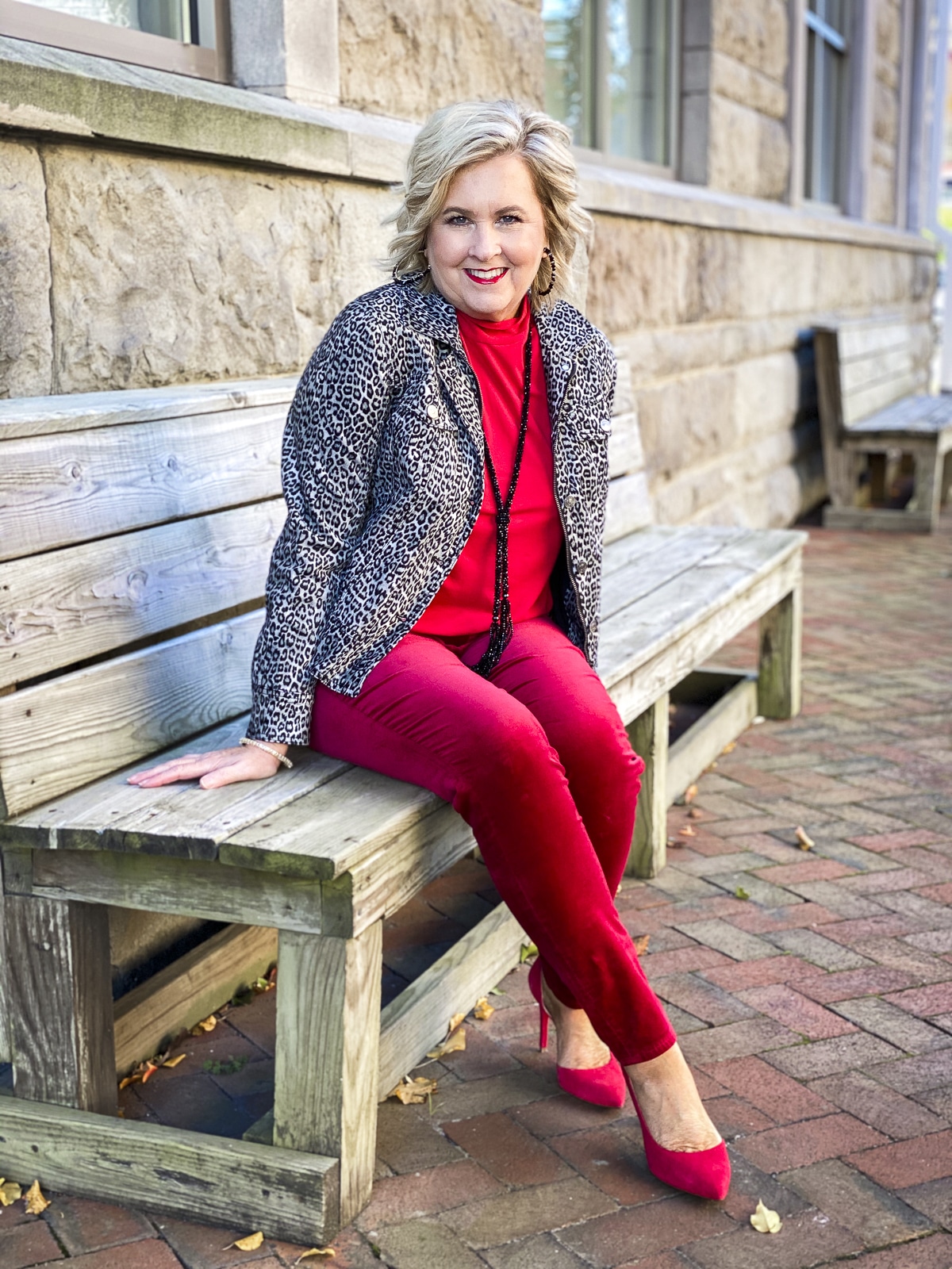 Fashion Blogger 50 Is Not Old is sitting on a bench and wearing a red outfit with a leopard velveteen jacket and black jewelry from Chico's