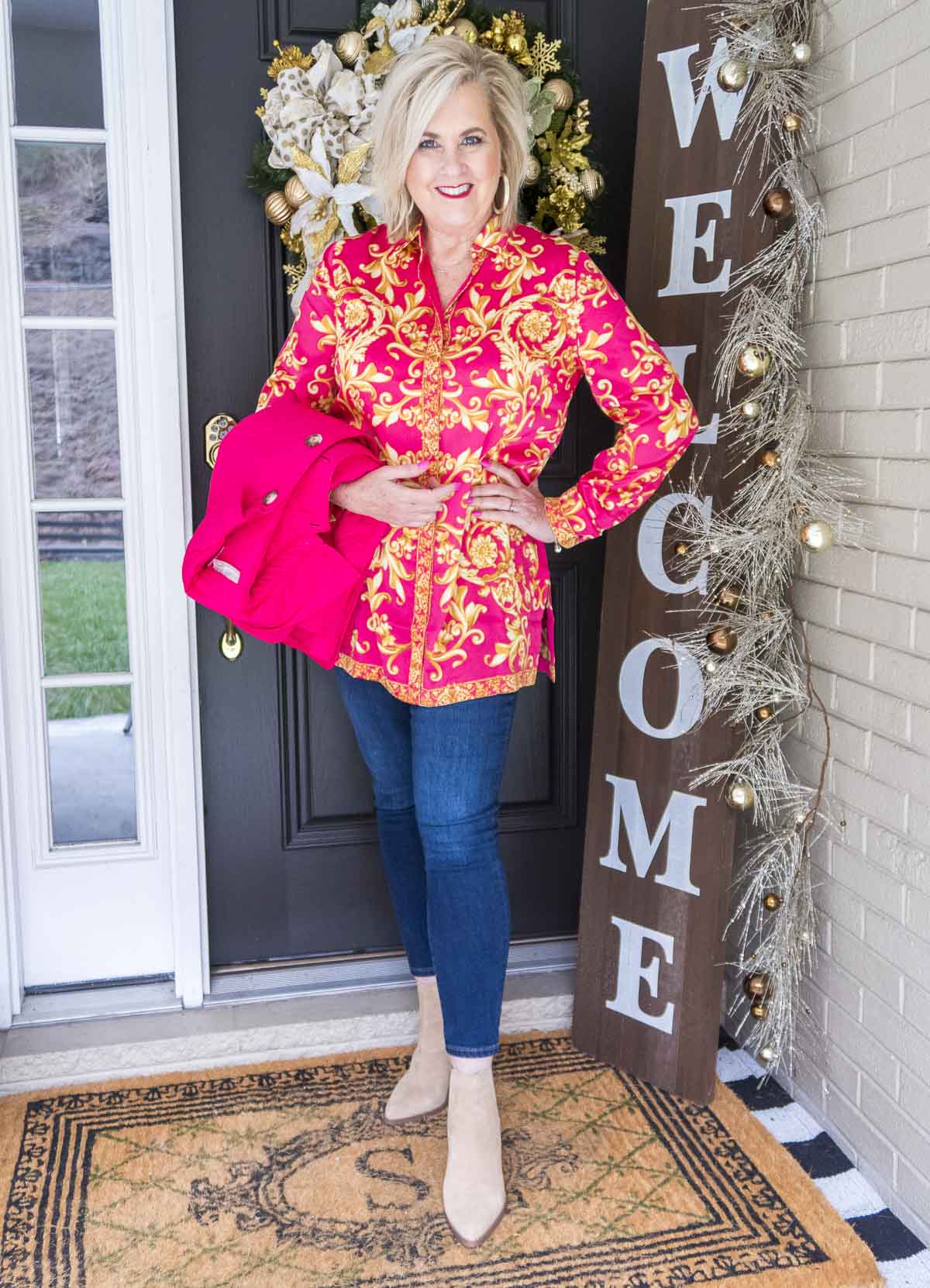 Fashion Blogger 50 Is Not Old is holding a brightly colored pink jacket and wearing a scroll print blouse with dark wash skinny ankle jeans and neutral colored booties