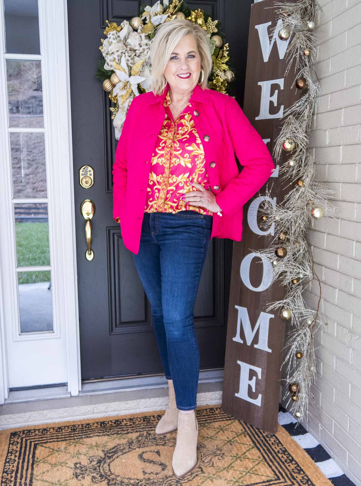 Fashion Blogger 50 Is Not Old is wearing a bright and bold colored pink jacket and scroll print blouse with dark skinny ankle jeans and neutral colored booties
