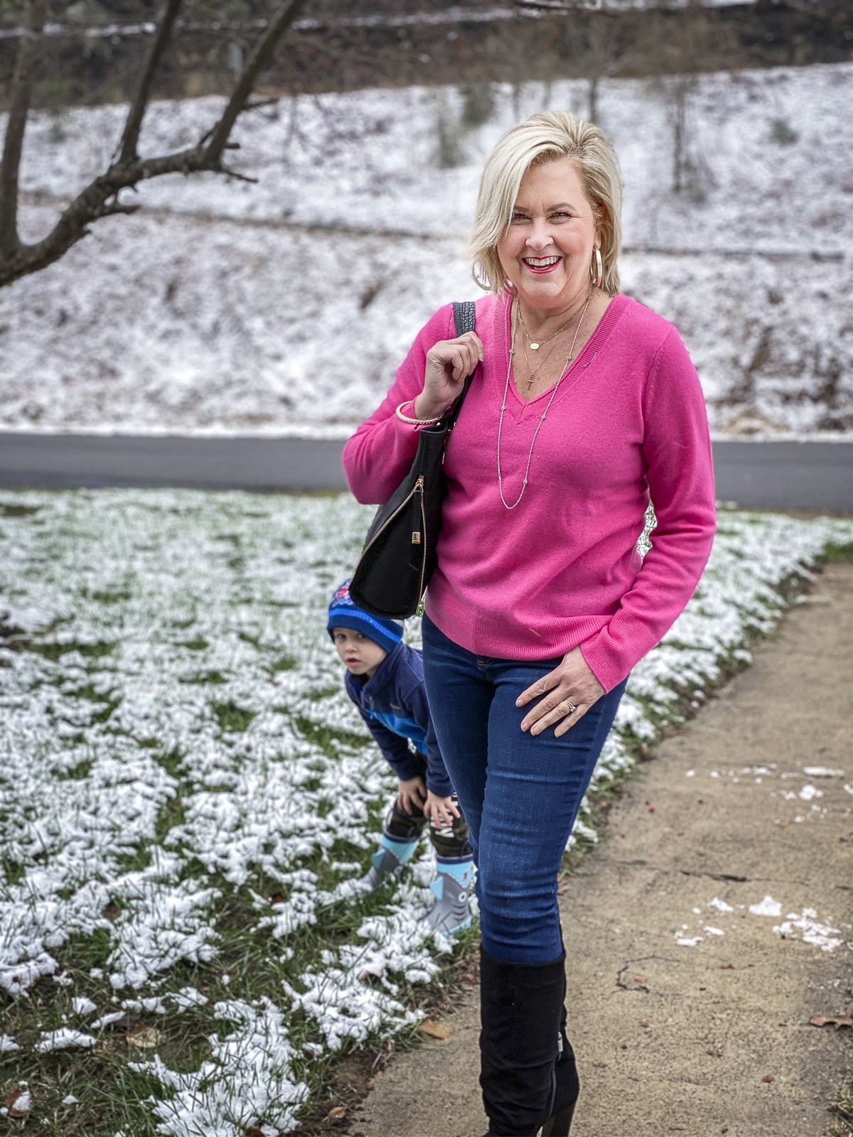 FASHION BLOGGER 50 IS NOT OLD is wearing a bright pink v-neck cashmere sweater, skinny jeans, and black suede boots while her grandson peeps around her legs