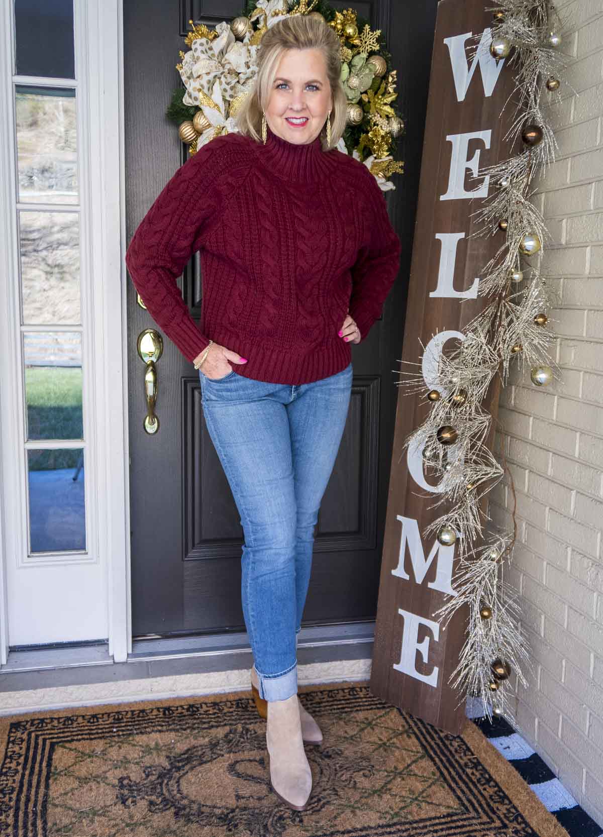 Fashion Blogger 50 Is Not Old is styling a cranberry cable-knit turtleneck sweater, a pair of denim jeans with a large cuff, and a pair of pointy toe ankle boots
