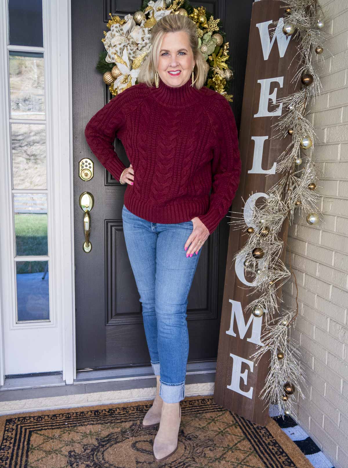 Fashion Blogger 50 Is Not Old is styling a cranberry cable-knit turtleneck sweater, a pair of denim jeans with a large cuff, and a pair of ankle boots