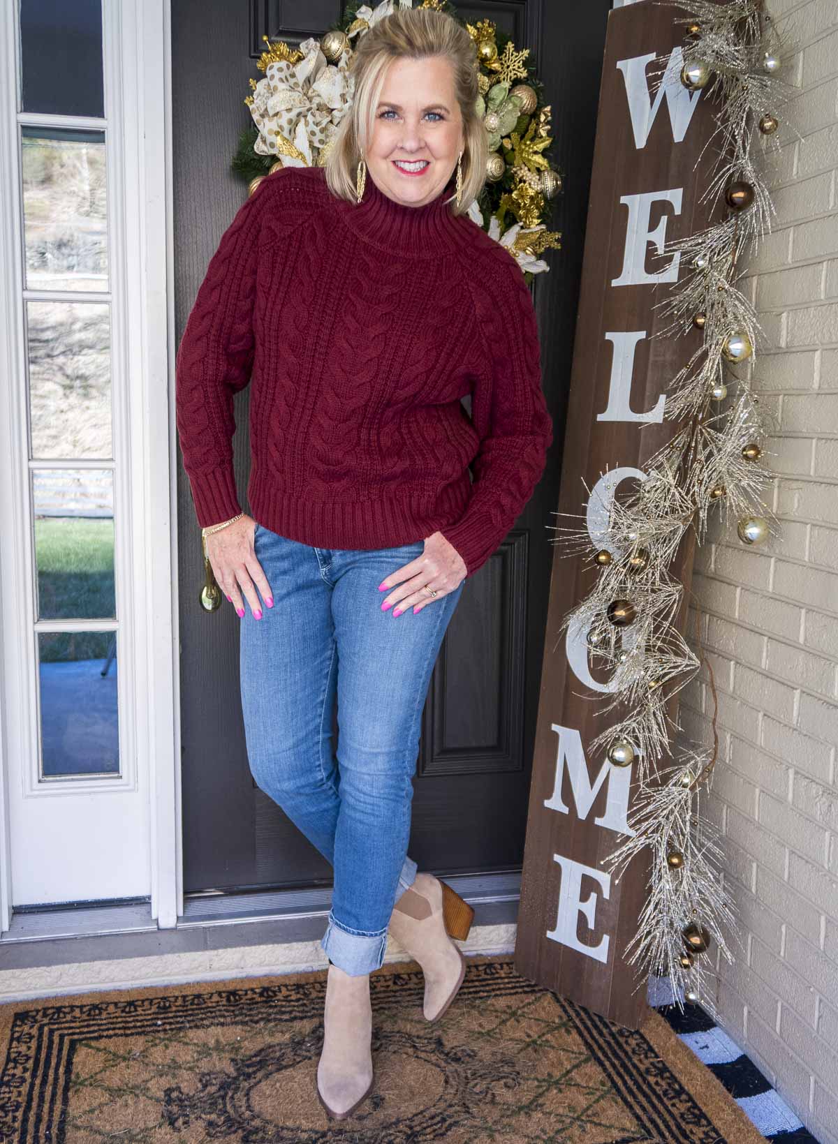 Fashion Blogger 50 Is Not Old is styling a cranberry cable-knit turtleneck sweater, a pair of denim jeans, and a pair of pointy toe ankle boots