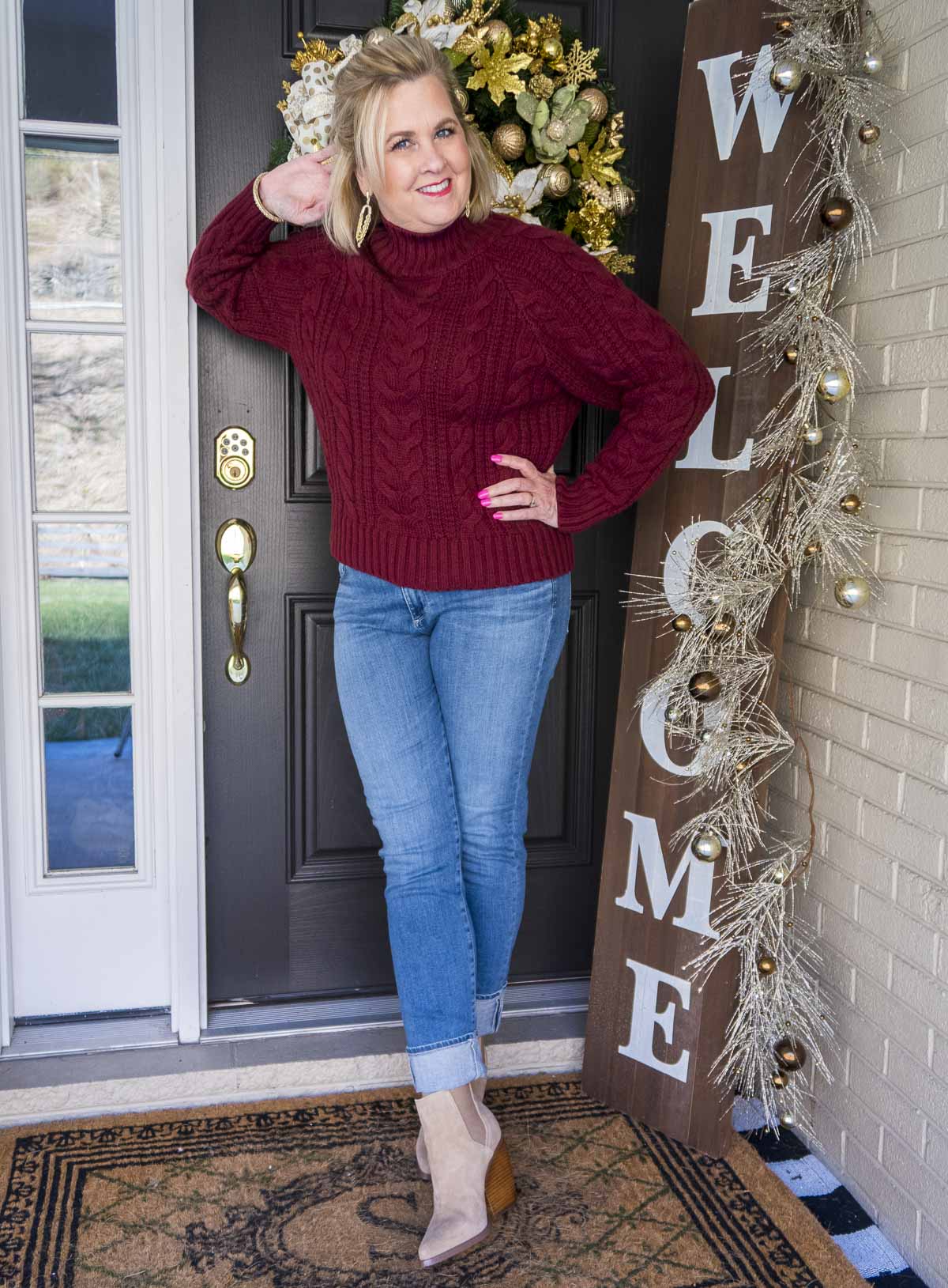 Fashion Blogger 50 Is Not Old is leaning on a door in a cranberry cable-knit turtleneck sweater, a pair of denim jeans with a large cuff, and a pair of pointy toe ankle boots