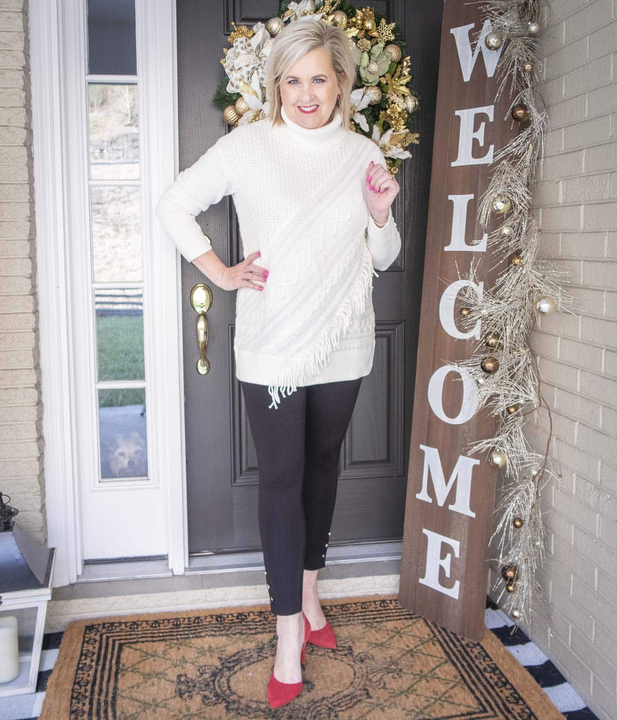 Fashion Blogger 50 Is Not Old is styling a Ecru sweater with unique details including fringe, a pair of black snap leggings, and red suede pumps