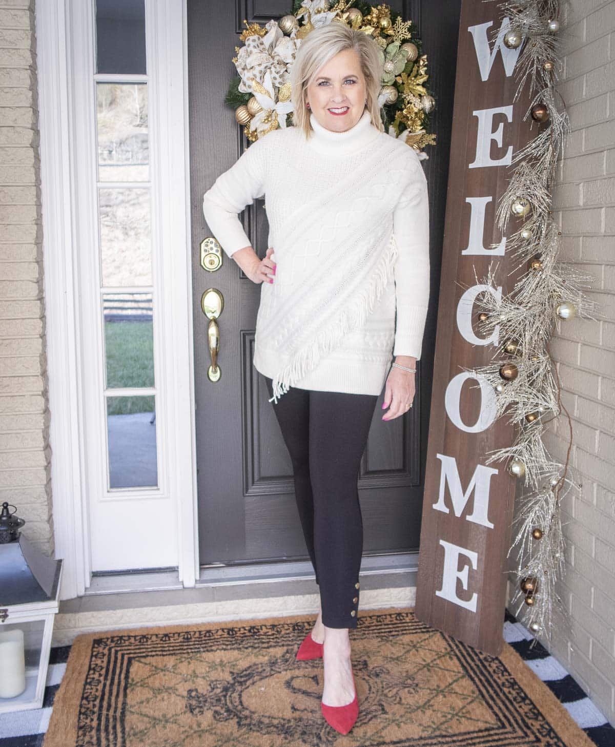 Fashion Blogger 50 Is Not Old is styling a Ecru sweater with unique details including fringe, a pair of black snap leggings, and red pumps