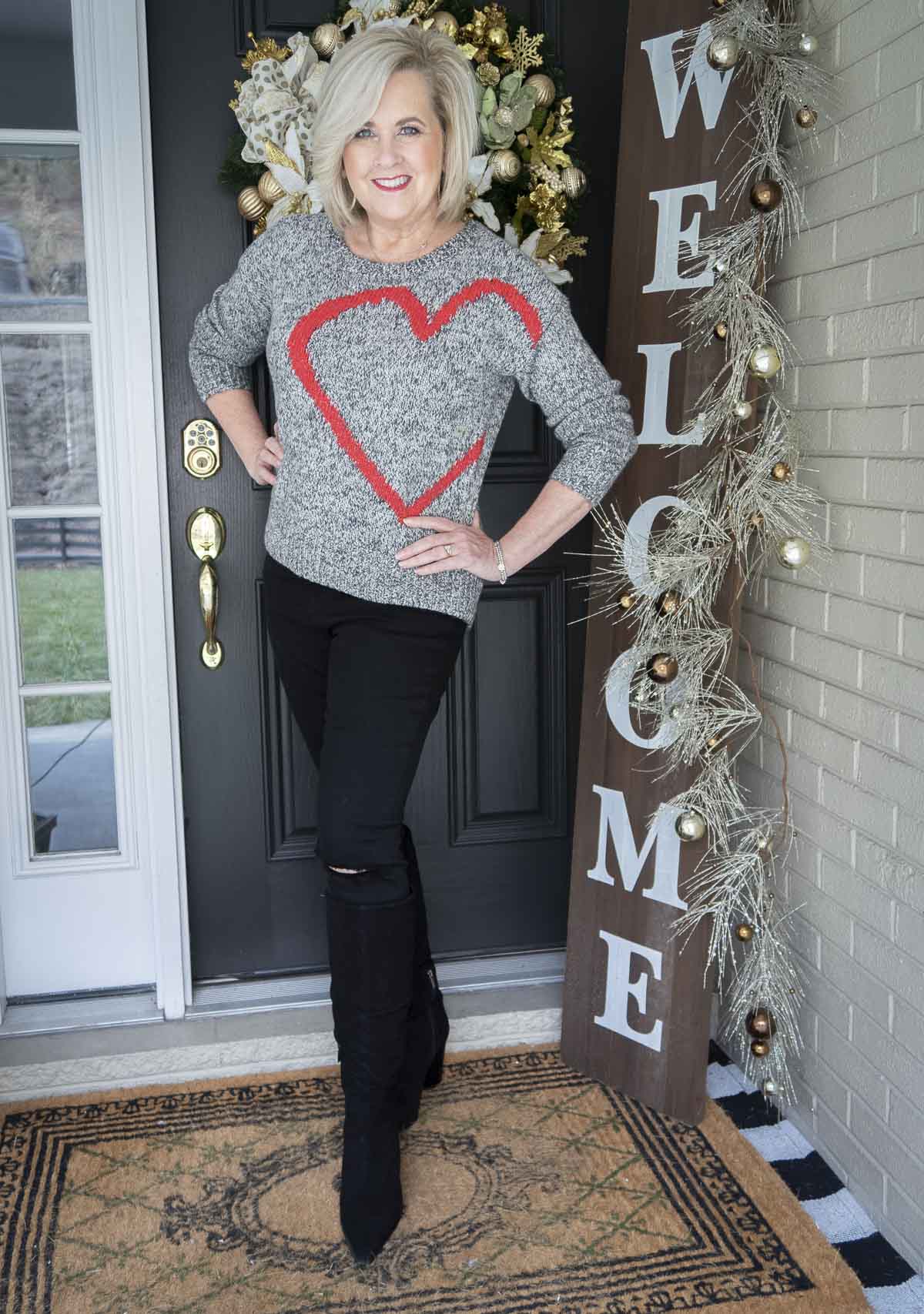 Fashion Blogger 50 Is Not Old is wearing a black and white sweater with a large red heart, a pair of black distressed jeans, and pointy toe black knee boots