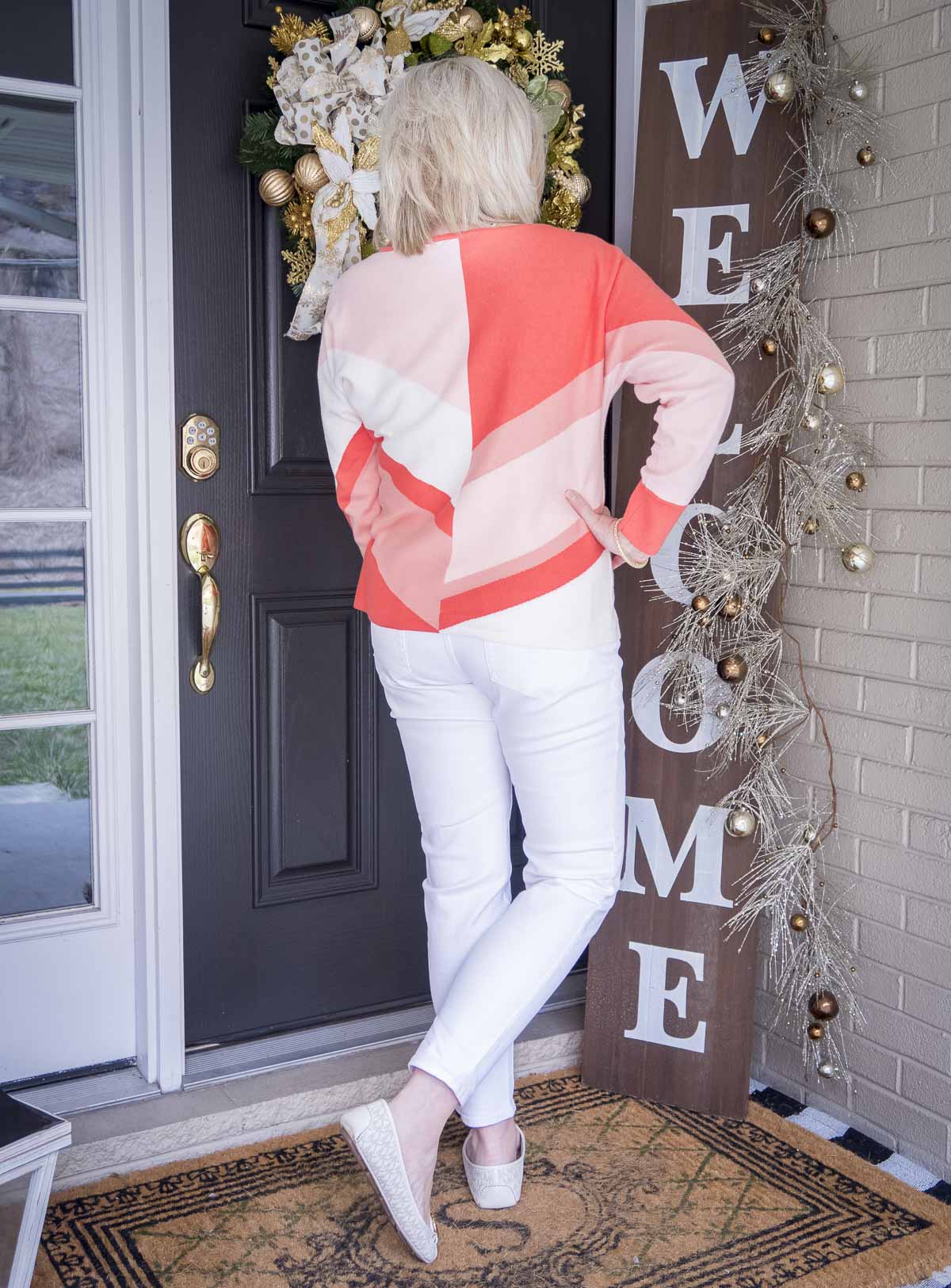 Fashion Blogger 50 Is Not Old is showing the back of a coral colorblock sweater, white ankle jeans, and white ballet flats for spring