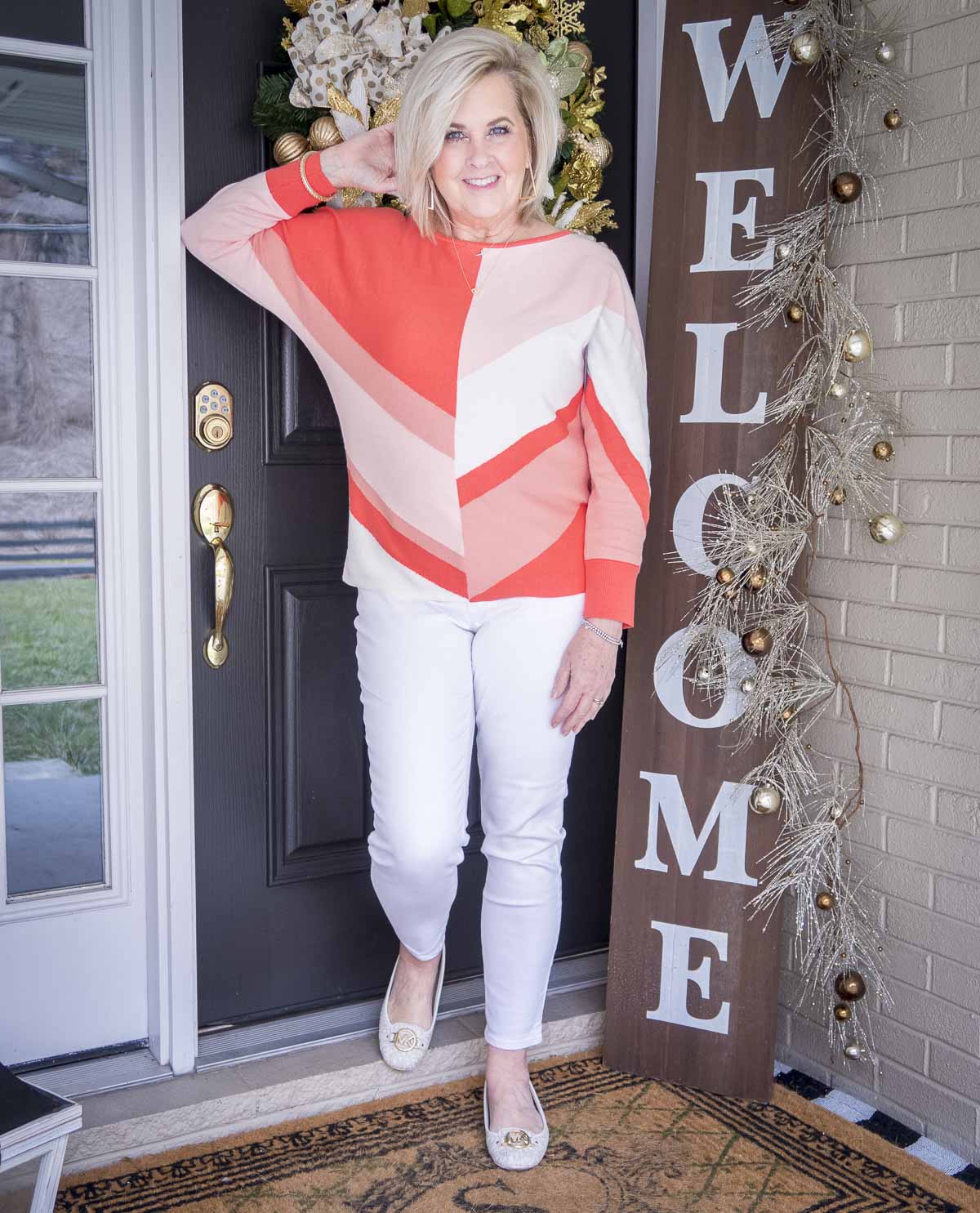 Fashion Blogger 50 Is Not Old is leaning against the door in a coral colorblock sweater, white ankle jeans, and white ballet flats for spring