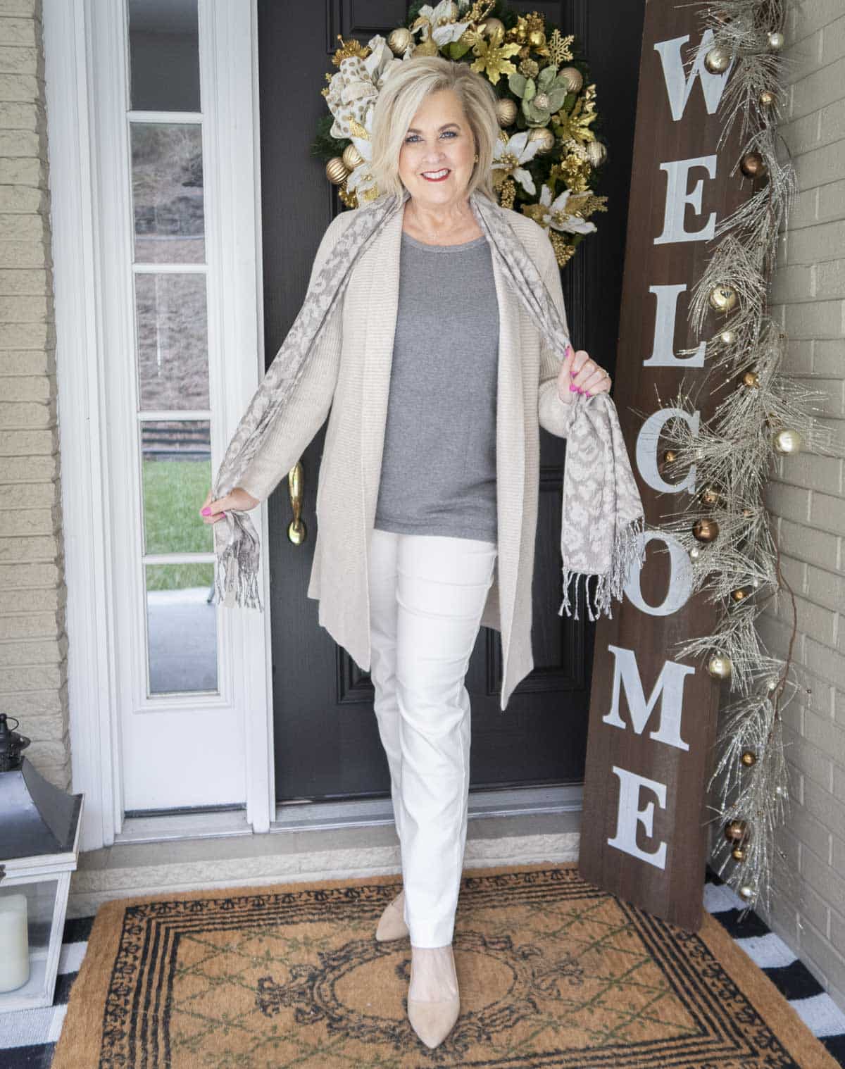 Fashion Blogger 50 Is Not Old is layering her outfit with a animal print scarf and a long neutral cardigan over a gray tank top and white pants