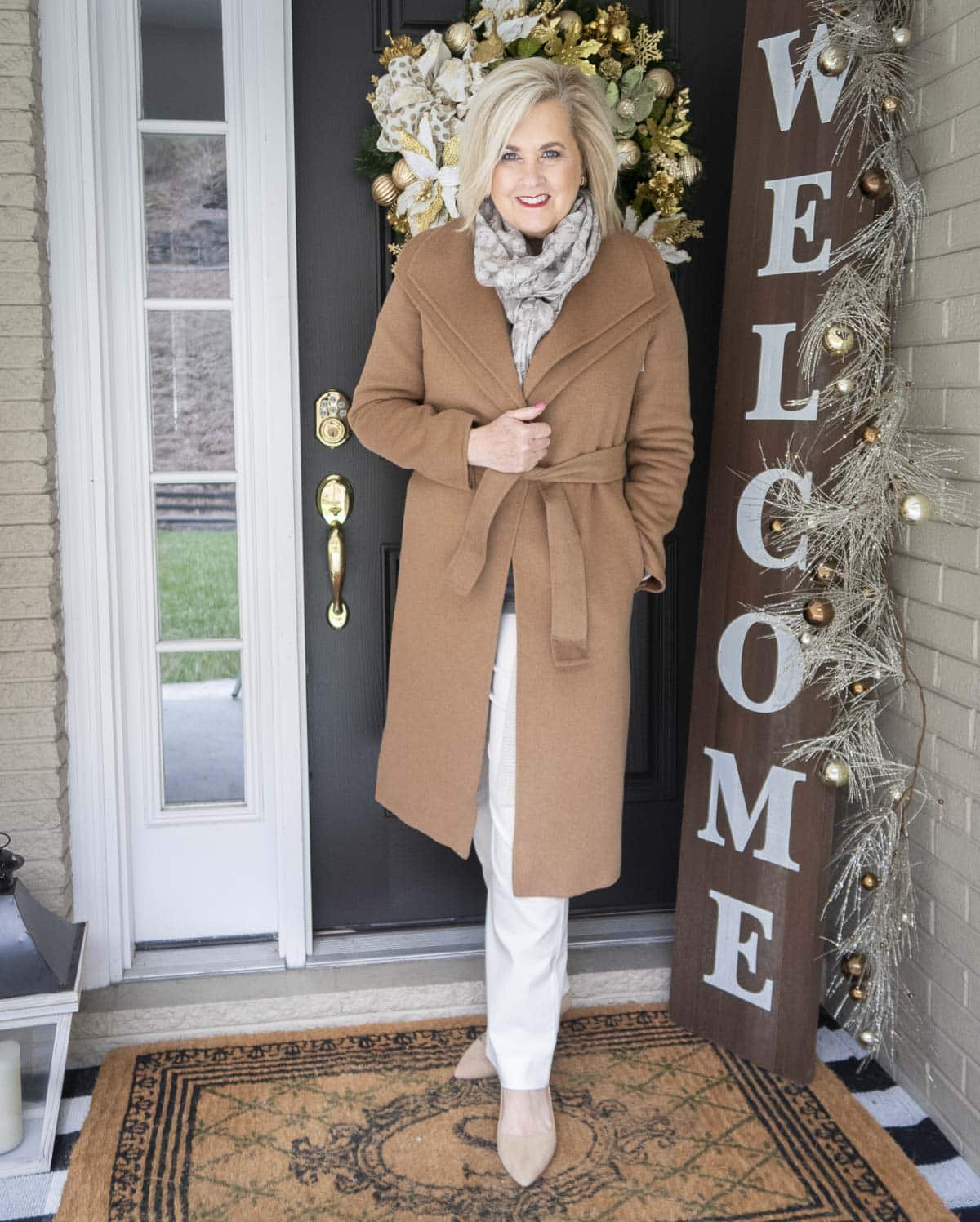 Fashion Blogger 50 Is Not Old is layering her outfit with a camel coat, an animal print scarf, and white pants