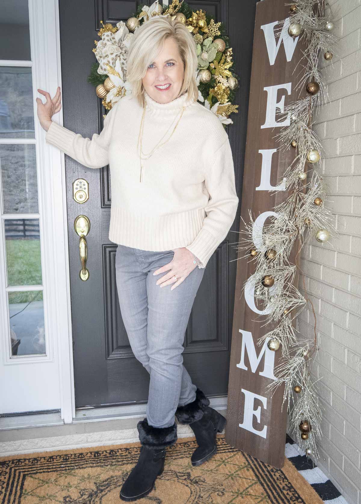 Fashion Blogger 50 Is Not Old is leaning against the door in an ivory turtleneck sweater, washed gray jeans, and black fur booties