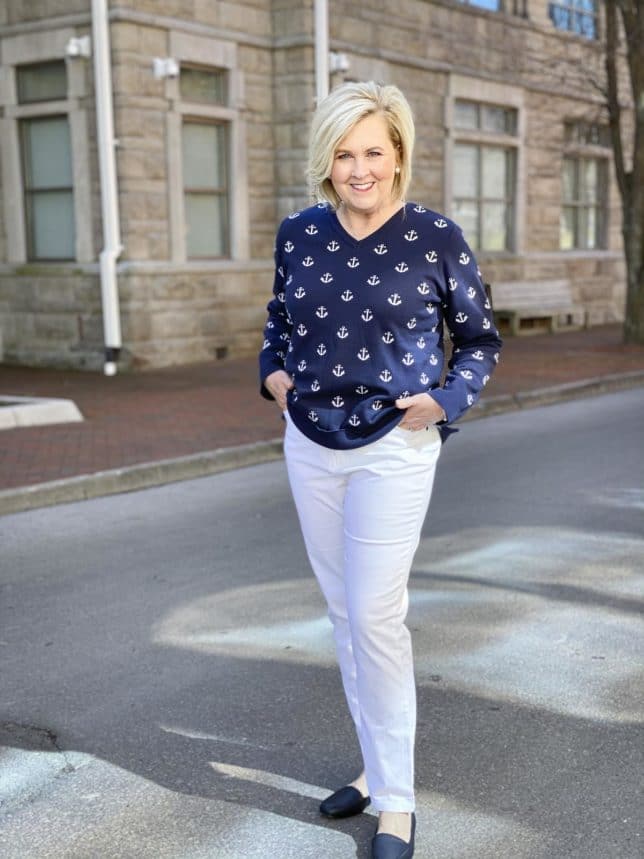 Fashion Blogger 50 Is Not Old is standing with her hands in her pockets wearing a navy sweaters with white anchors, white ankle pants, and navy flats