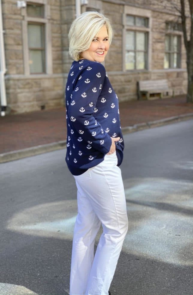 Fashion Blogger 50 Is Not Old is looking over her shoulder in a navy sweaters with white anchors, white ankle pants, and navy flats