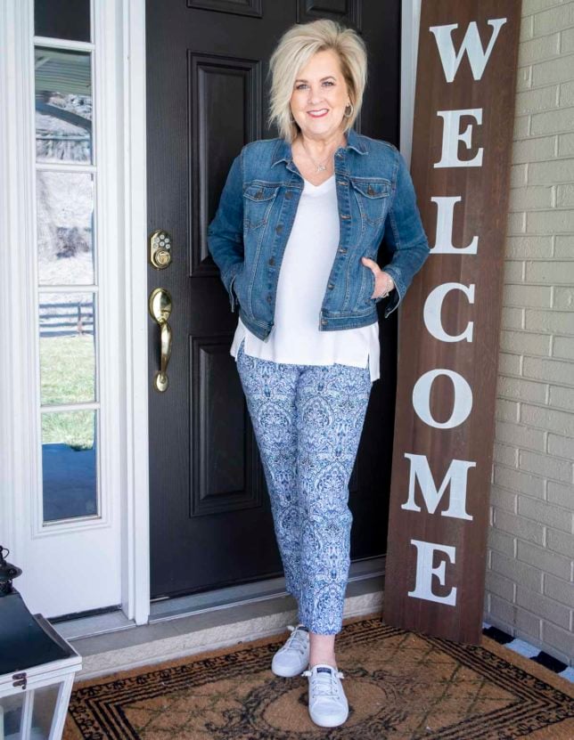 Fashion Blogger 50 Is Not Old is wearing a white v-neck tunic, a classic denim jacket, a blue printed ankle pant, and white Sperry sneakers