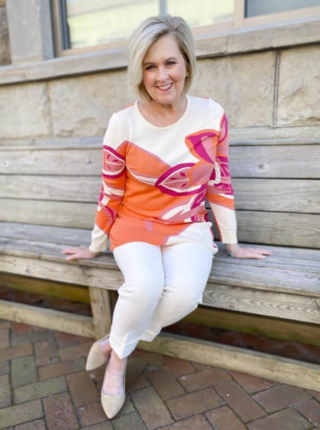 Fashion Blogger 50 Is Not Old is sitting on a bench in a tunic sweater with a large floral print, ecru cuffed dress pants, and neutral kitten heels