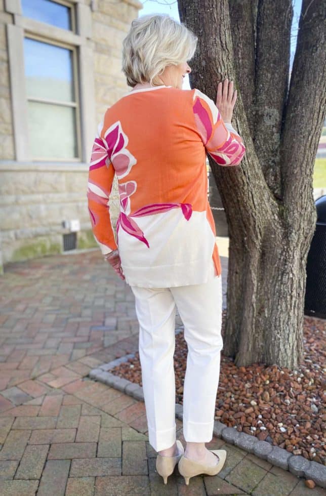 Fashion Blogger 50 Is Not Old is showing the back of a beautiful tunic sweater with a large floral print, ecru cuffed dress pants, and neutral kitten heels