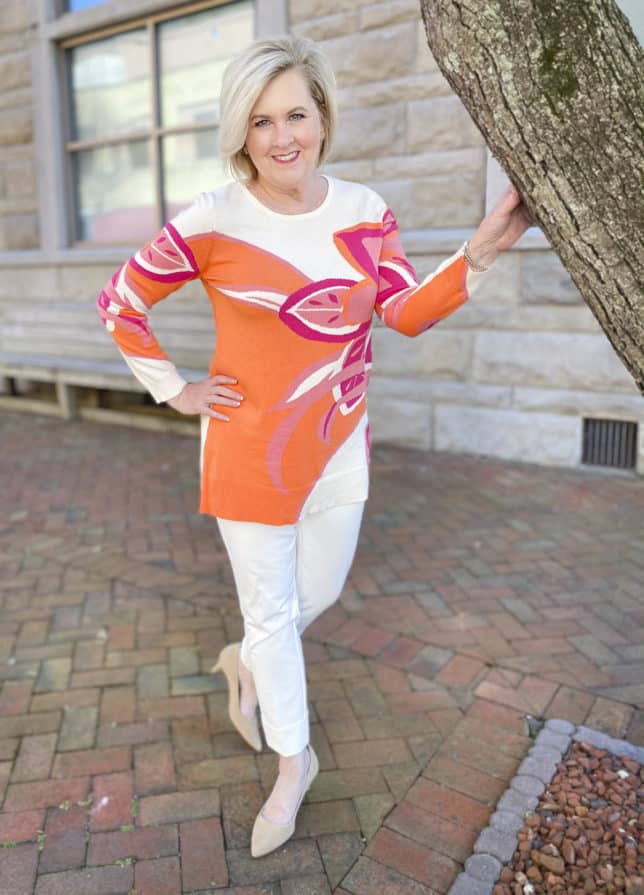Fashion Blogger 50 Is Not Old is wearing a tunic sweater from Chicos with a large floral print, ecru cuffed dress pants, and neutral kitten heels