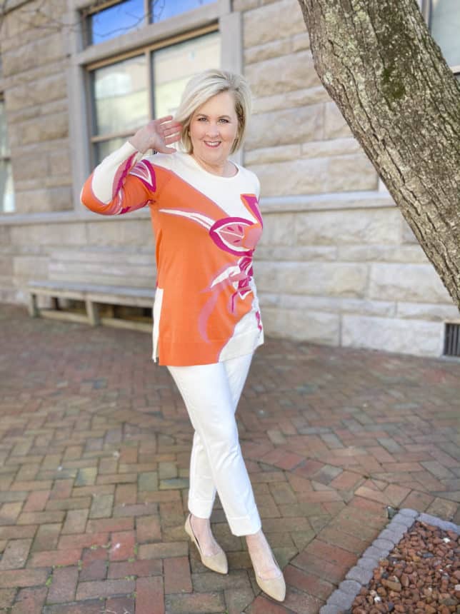 Fashion Blogger 50 Is Not Old is moving the hair from her face while wearing a beautiful tunic sweater with a large floral print, ecru cuffed dress pants, and neutral kitten heels