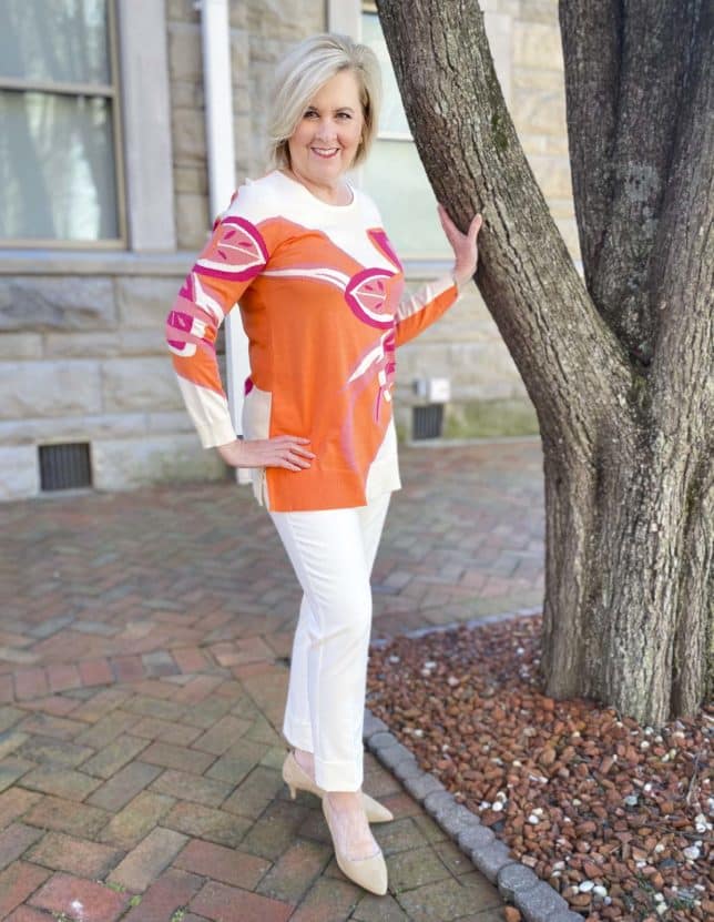 Fashion Blogger 50 Is Not Old is standing near a tree wearing a beautiful tunic sweater with a large floral print, ecru cuffed dress pants, and neutral kitten heels
