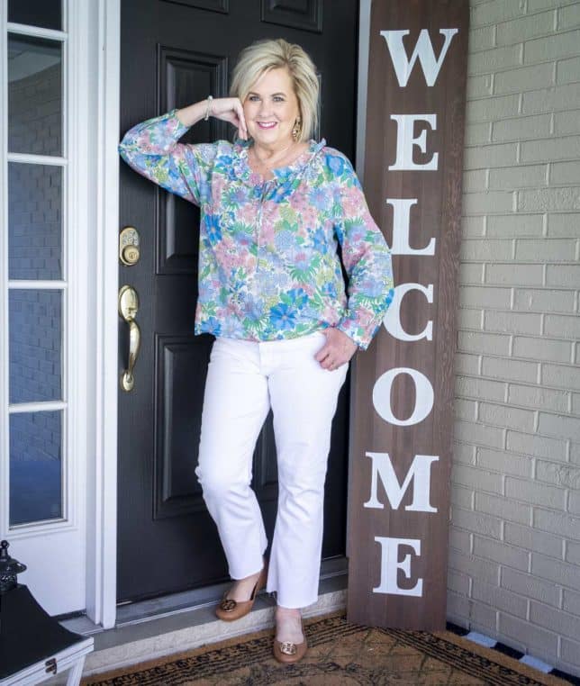 Fashion Blogger 50 Is Not Old is leaning against a door wearing white cropped jeans with a floral top, and Tory Burch ballet flats