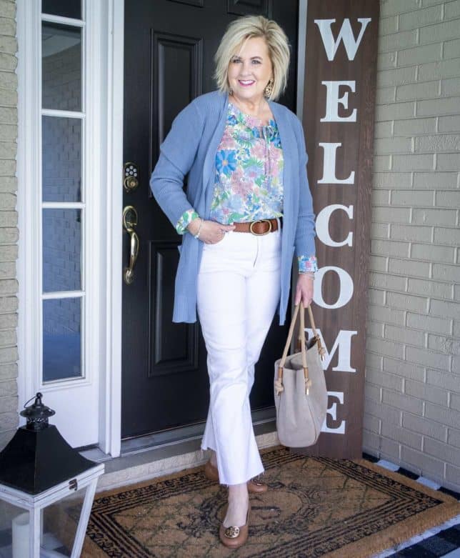 Fashion Blogger 50 Is Not Old is wearing white cropped jeans with a floral top, a long blue cardigan, and a brown belt with a Tory Burch ballet flats